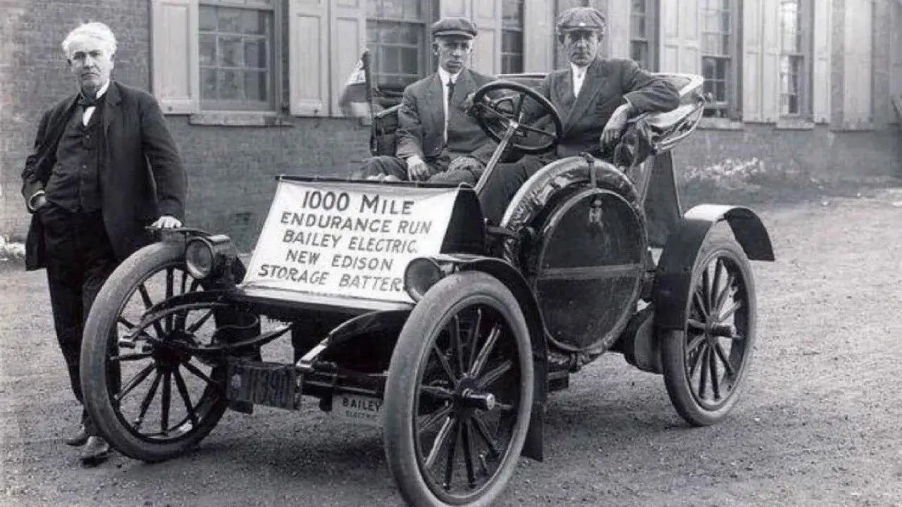 Un hombre junto a un coche eléctrico Bailey de 1907, parte de la historia del coche eléctrico, con un cartel que anuncia una carrera de resistencia de 1000 millas.