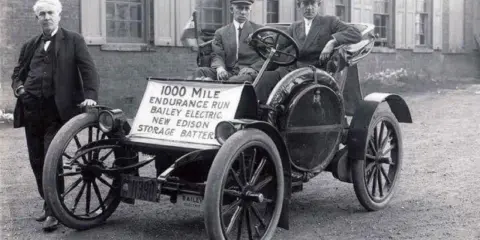 Un hombre junto a un coche eléctrico Bailey de 1907, parte de la historia del coche eléctrico, con un cartel que anuncia una carrera de resistencia de 1000 millas.