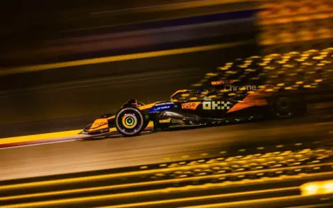 Un coche de **fórmula 1** naranja y negro acelera en la noche, dejando estelas de luz dorada.