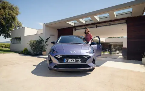 Mujer sonriente junto a un coche alto y morado, aparcado frente a una casa moderna con puertas abiertas.