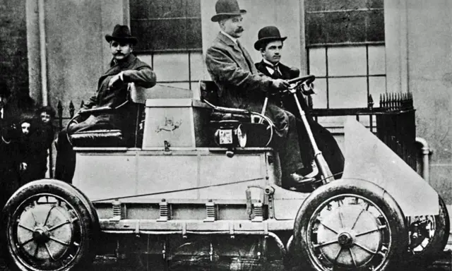 Hombres con sombreros posan en el primer coche eléctrico de la historia, un vehículo pionero de finales del siglo XIX.
