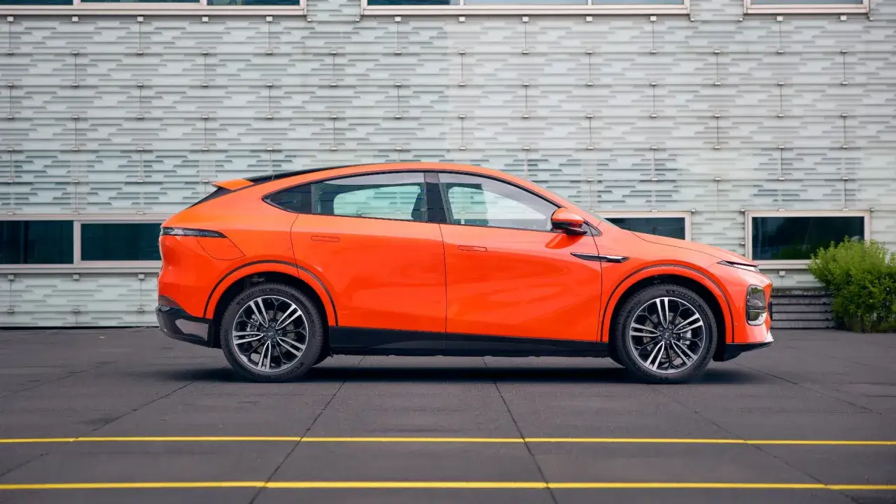 Un elegante coche el&eacute;ctrico grande de color naranja brillante, con l&iacute;neas deportivas y un dise&ntilde;o moderno, aparcado frente a un edificio con fachada texturizada.