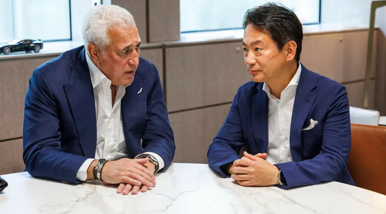 Dos hombres de negocios, uno de ellos Koji Watanabe, conversan sentados a una mesa. Detr&aacute;s, un coche en miniatura.